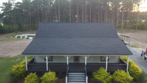 Aerial view of single-story house with dark shingle roof and white porch surrounded by forest