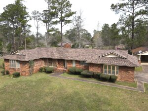 Red brick ranch style house with shingle roof surrounded by mature trees and lawn
