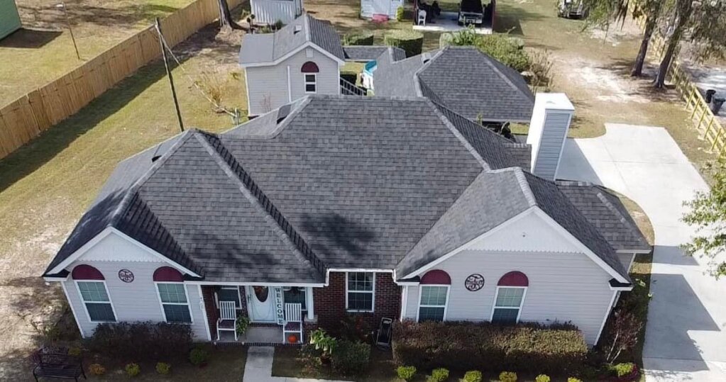 Aerial view of single-story white brick home with gray shingle roof and landscaping