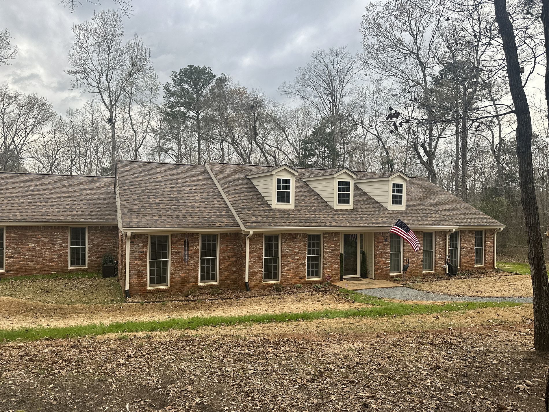 Single-story brick ranch house with dormer windows and American flag in wooded setting