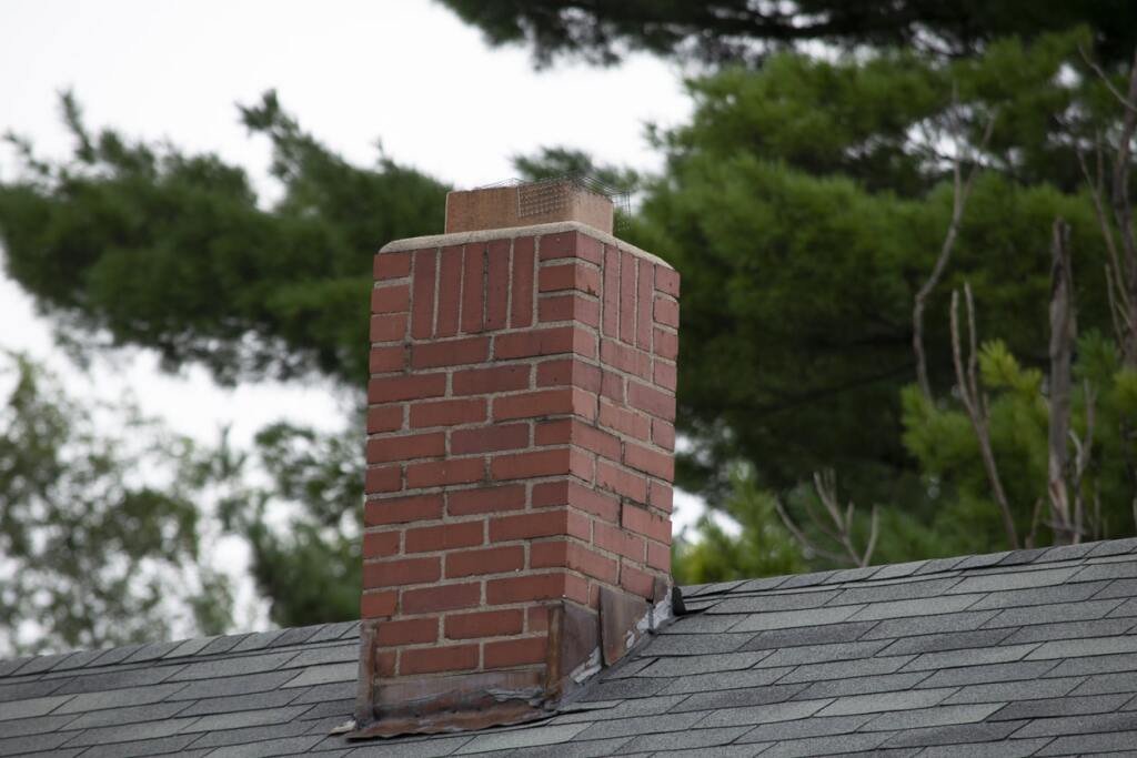 Red brick chimney on asphalt shingle roof surrounded by green trees