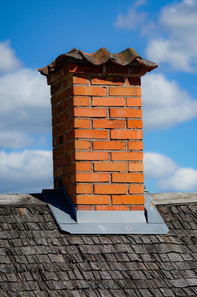 Red brick chimney with clay tile cap on wooden shingle roof against blue sky