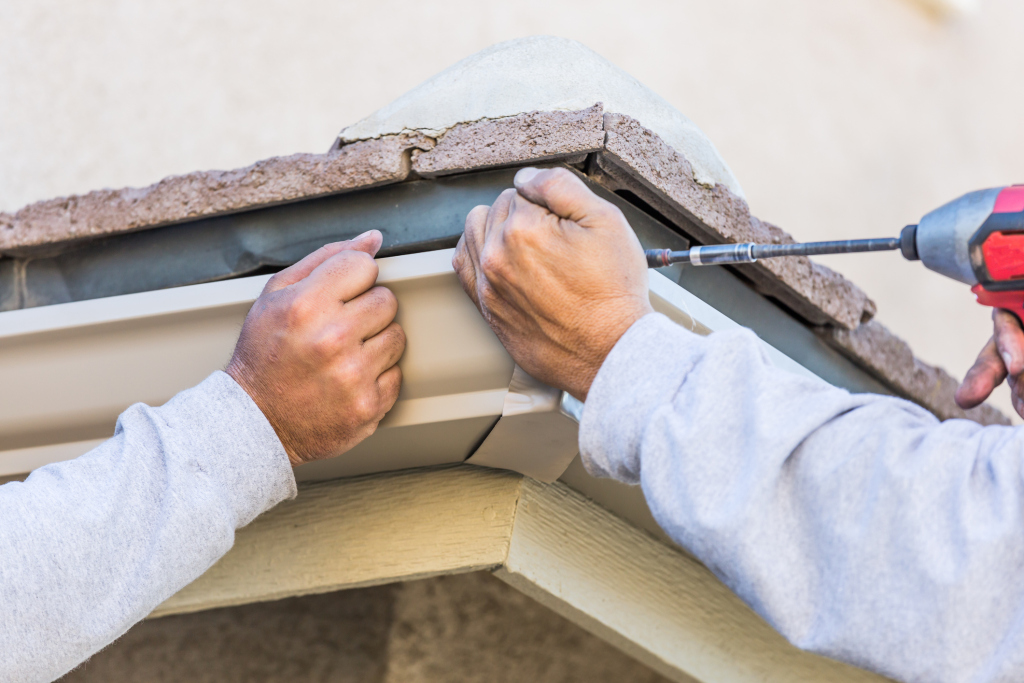 Worker installing white rain gutter with drill on residential home exterior