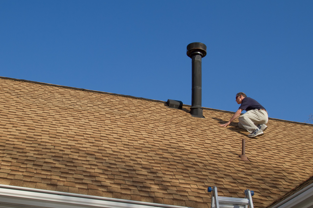Roofer working on asphalt shingle roof near black chimney pipe against blue sky