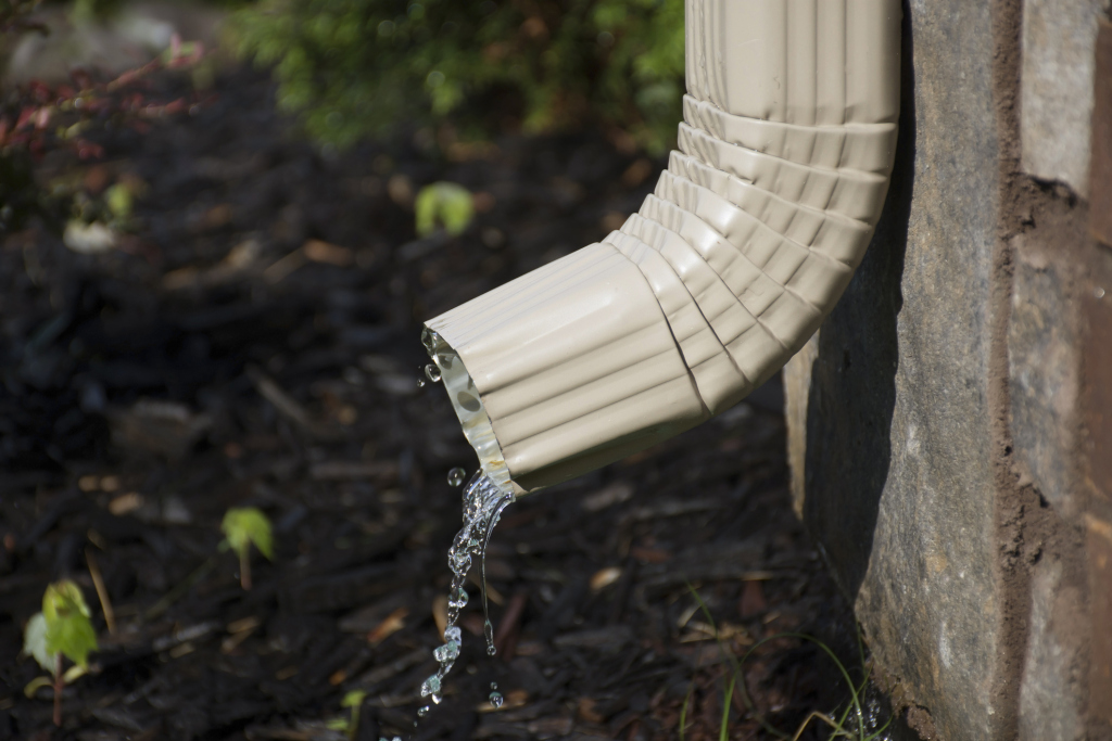 White rain gutter downspout with water flowing out onto garden ground