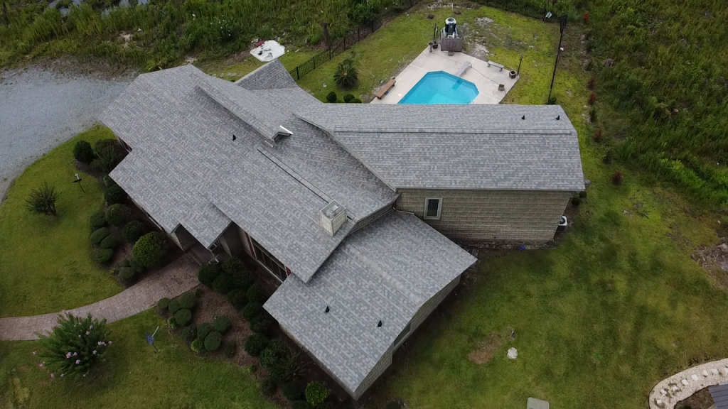 Aerial view of large suburban house with gray shingle roof and backyard pool