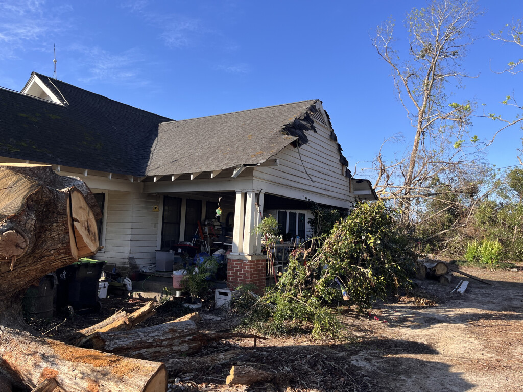 Storm damaged house with torn roof shingles and fallen tree debris in yard