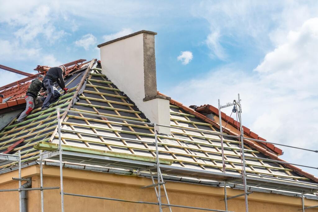 Roofers installing red clay tiles on house roof with scaffolding and chimney
