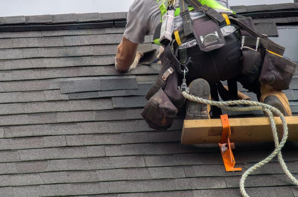 Roofer in safety harness installing shingles on residential roof with professional tools