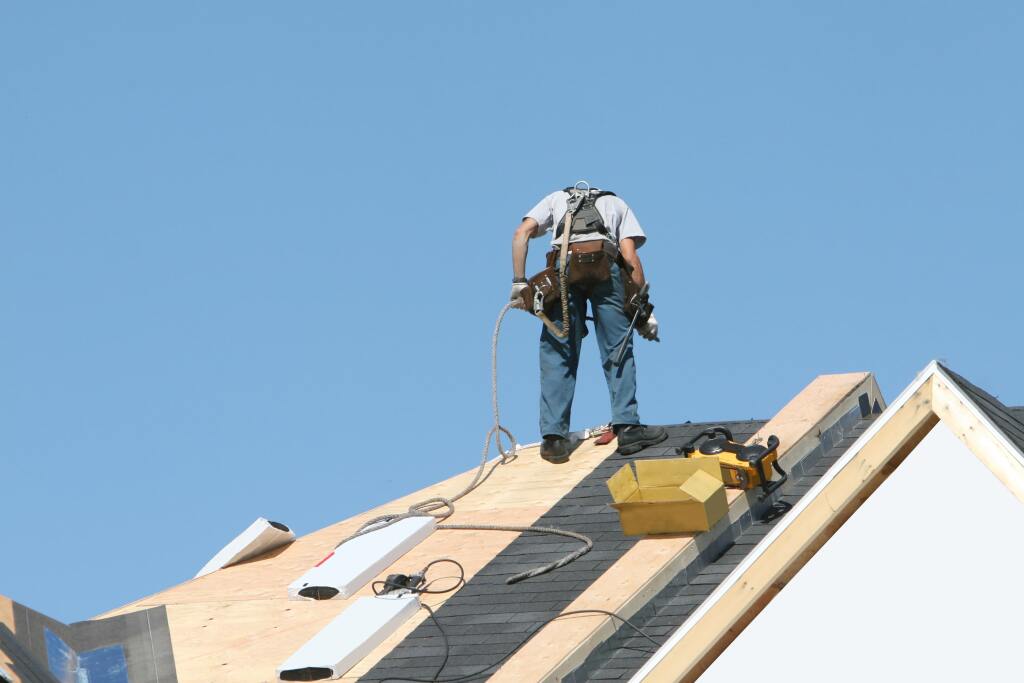Roofer with safety harness installing shingles on residential roof against blue sky